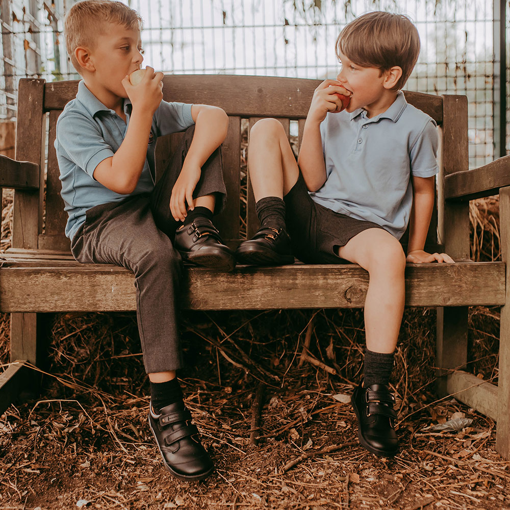 Two young boys in bobux shoes, sat on a wooden bench eating an apple