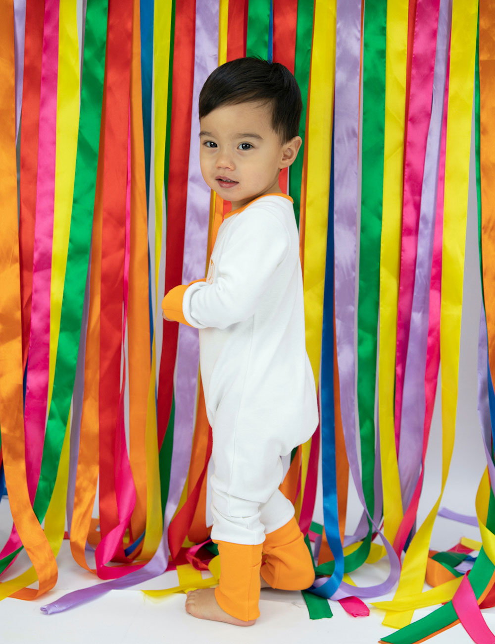 Dark haired boy in white sleep suit next to bright coloured ribbons