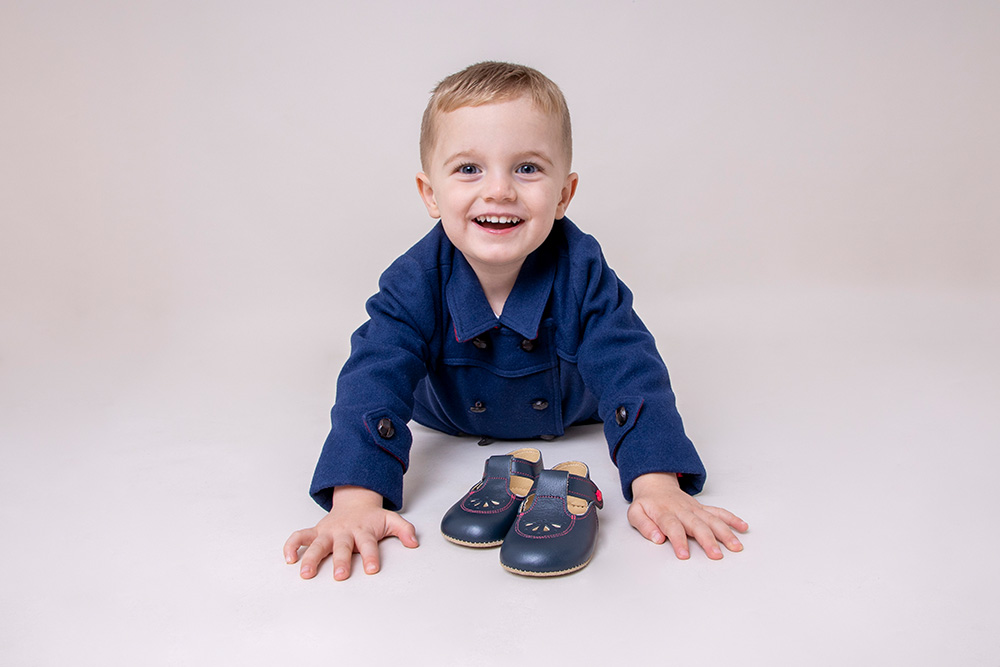 A young boy in a dark blue jacket laid on the floor in front of a pair of shoes