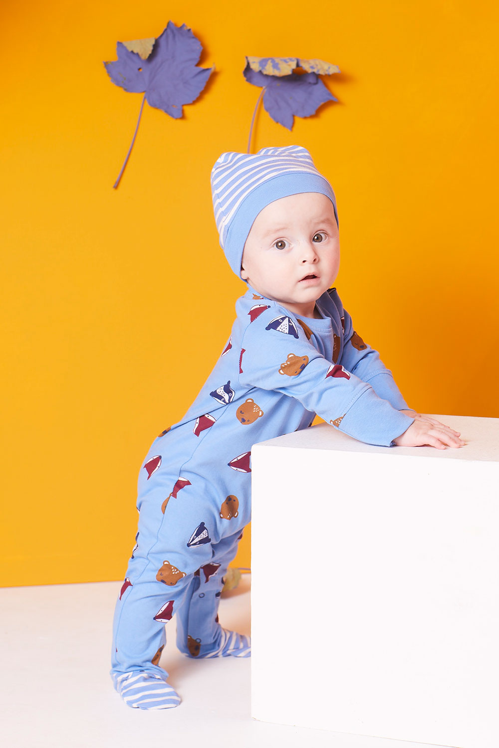 A young boy stood up wearing a blue baby grow by Lilly + Sid