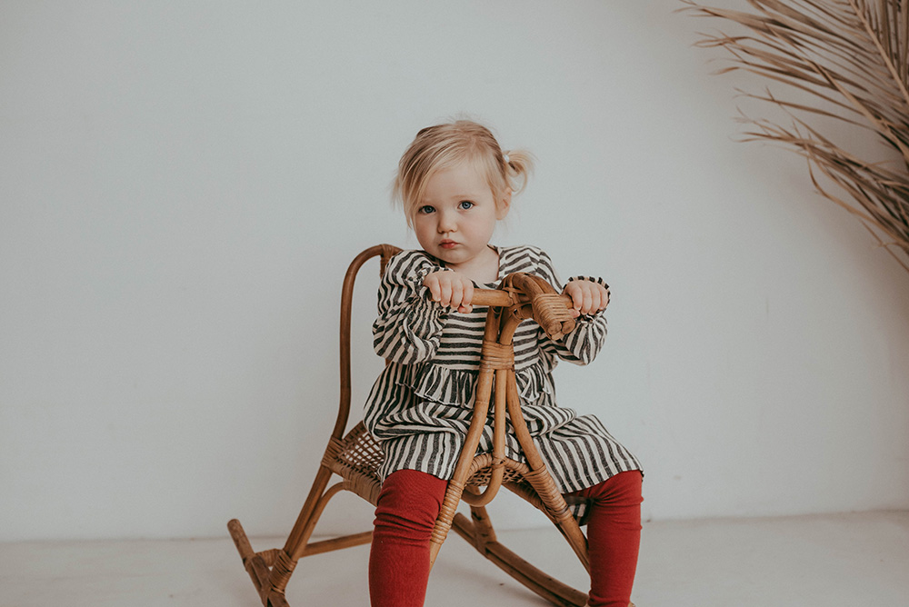 A young girl sat on a wooden rocking chair wearing a striped dress