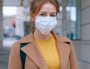 Woman in face mask wearing camel coloured coat and yellow jumper