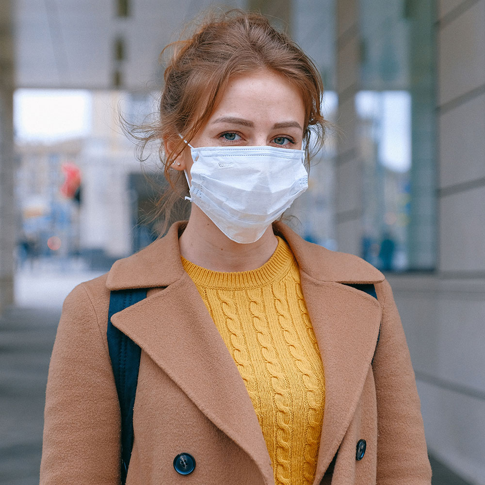 Woman in face mask wearing camel coloured coat and yellow jumper