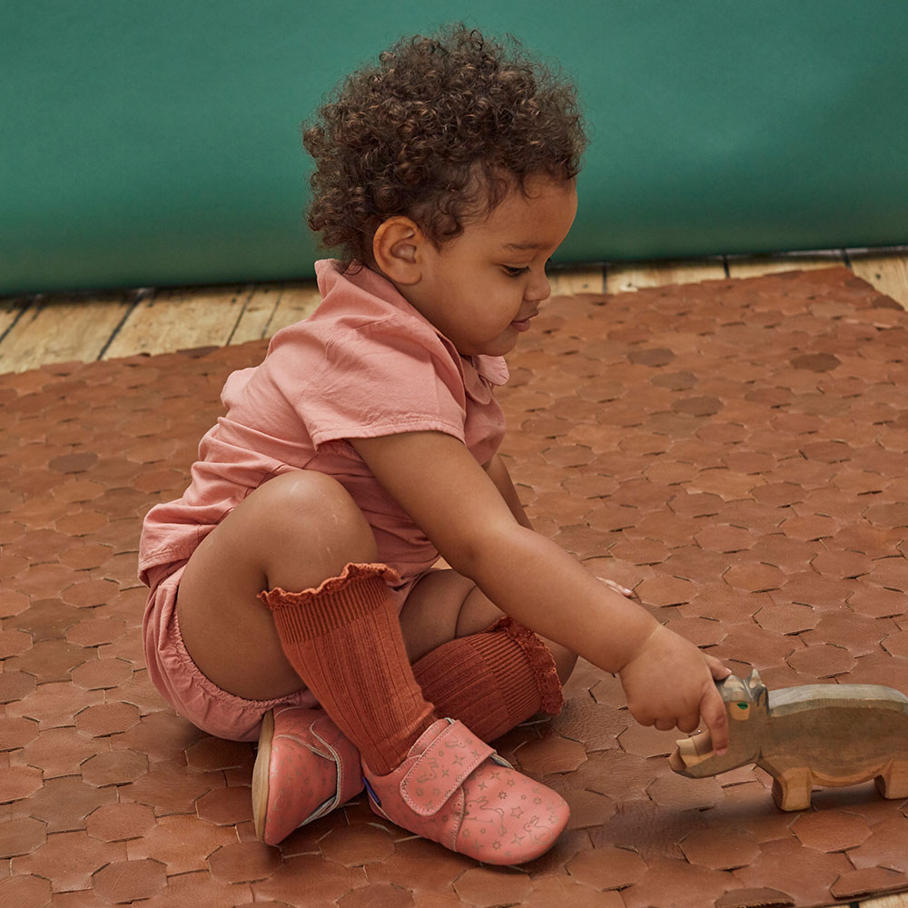 A young girl sat on the floor playing with a wooden rhino toy and wearing Poco Nido shoes