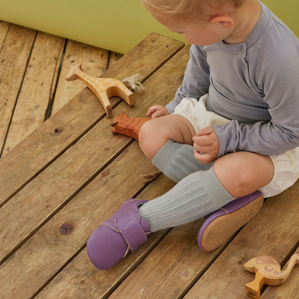 A young boy sat on the floor wearing purple velco strap Poco Nido shoes