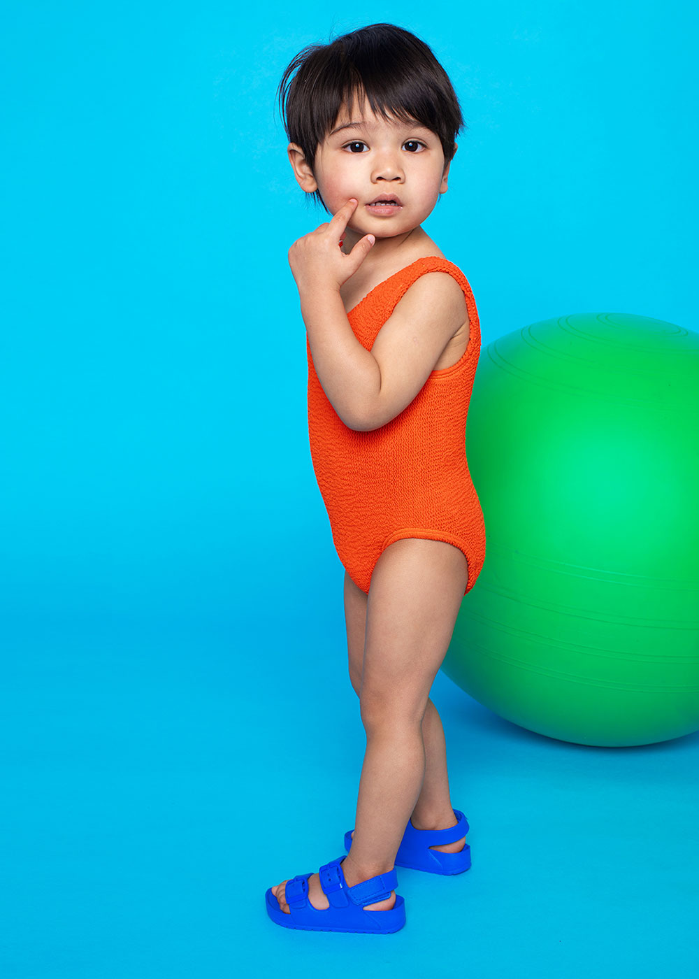 Young boy on bright orange Mini Maxx swimwear stood against bright blue background