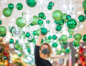 Woman in mask hanging bright green christmas baubles from the ceiling