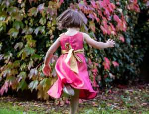 Girl running towards trees in pink dress with gold ribbon by Pri Pri