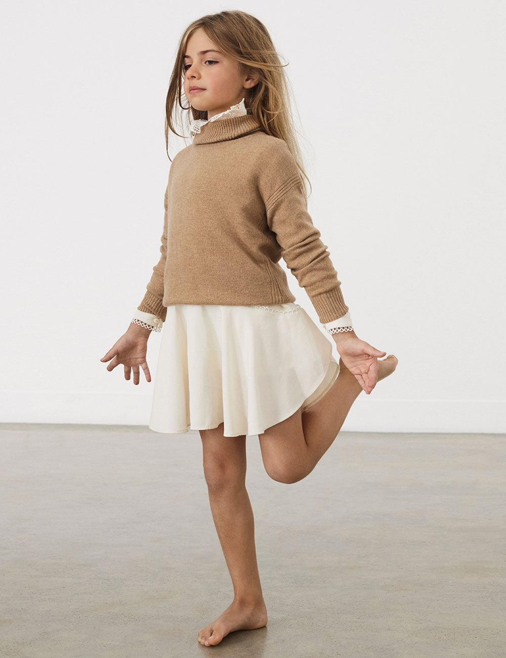 Young girl stood on one leg wearing beige Reiss jumper and white skirt
