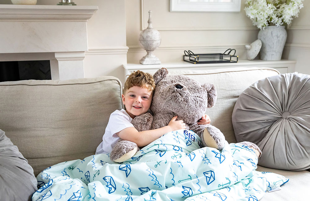 Little boy holding a grey teddy bear and underneath a blue blanket