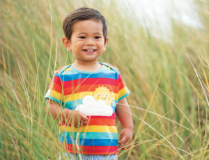 Young boy standing in long green grass wearing colourful striped Frugi T Shirt