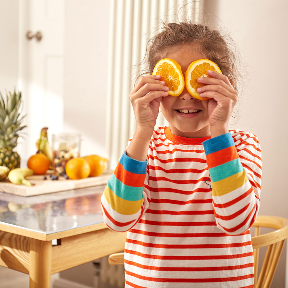 Girl wearing colourful striped Frugi top holding slives of orange to her eyes