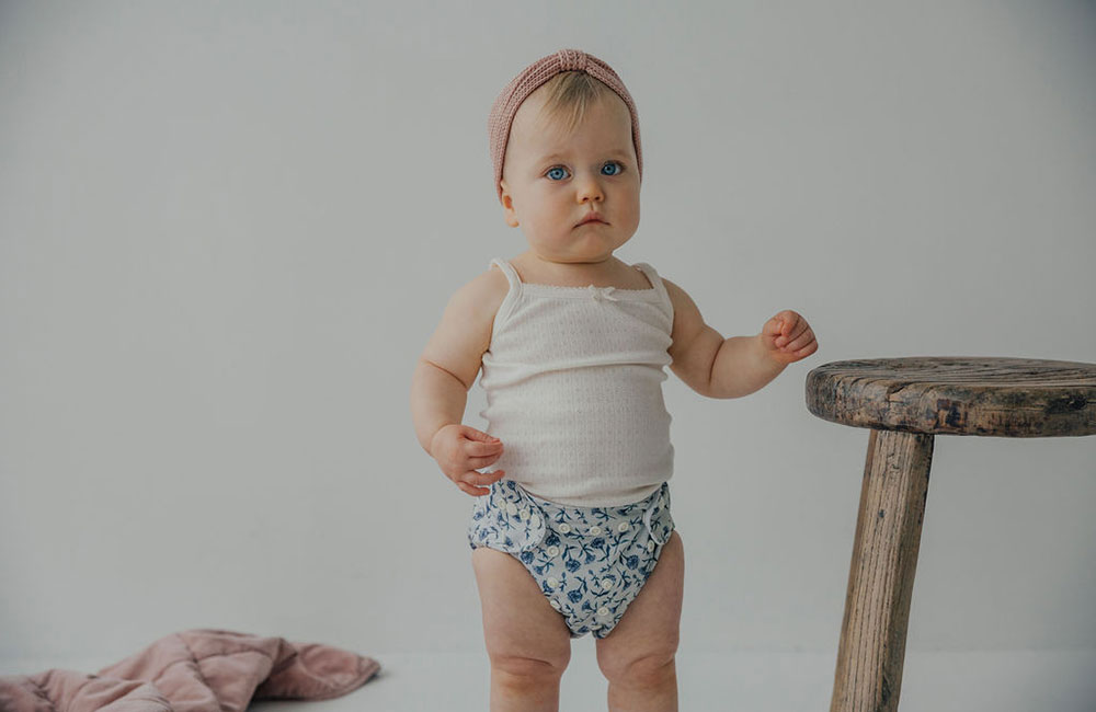 Baby girl stood up wearing white vest and blue reusable nappy