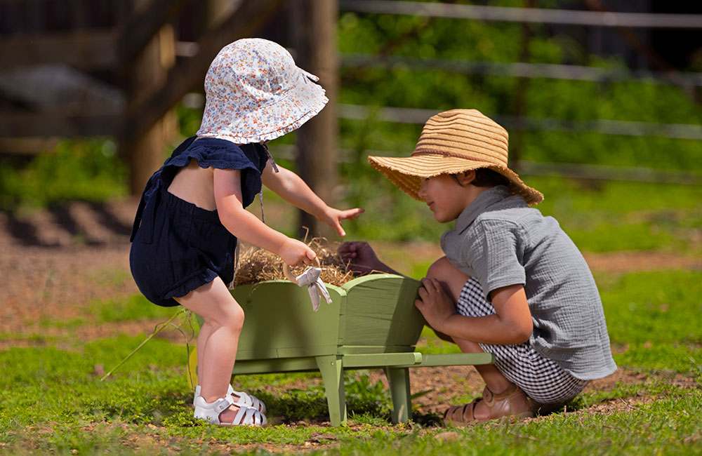Boy and girl playing in the grass with small green Bobux gardening wheelbarrow