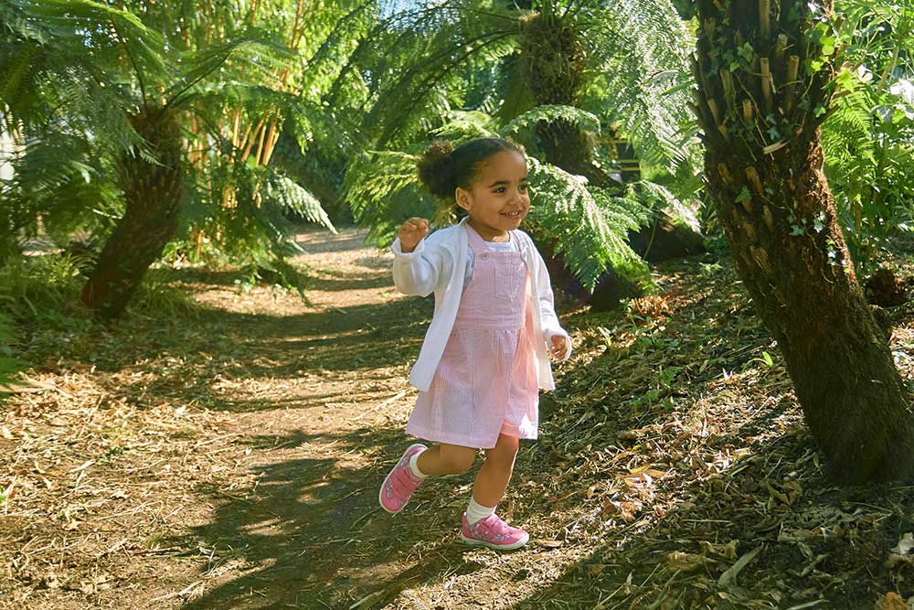 A young girl in a pink dress running through woodland wearing shoes by Start-Rite and Jojo Maman Bebe