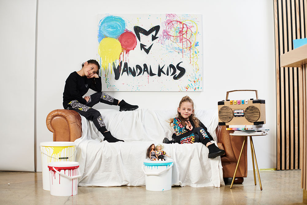 Two young girls sat on a sofa with a Vandal Kids logo painting on the wall behind them