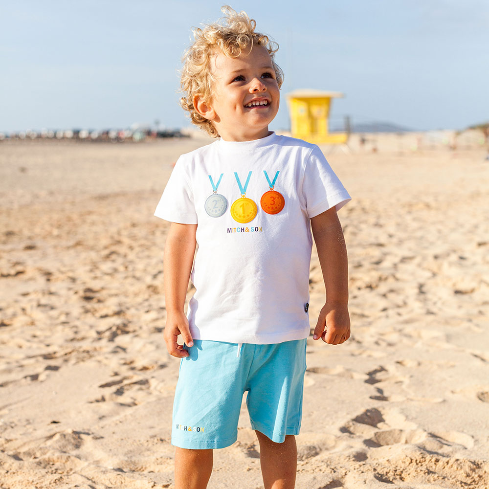 Boy stood on beach in shorts and T-shirt by Picture Book Fashion