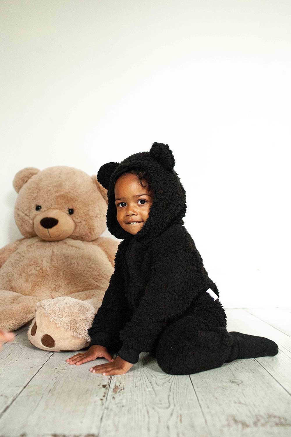 A young child sat on the floor next to a large teddy bear wearing a black bodysuit