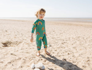 A young boy stood on the beach wearing UV protective swimwear by Muddy Puddles