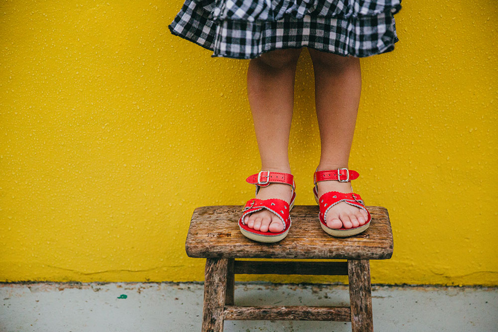 A young girl stood on a wooden stool wearing red sandals