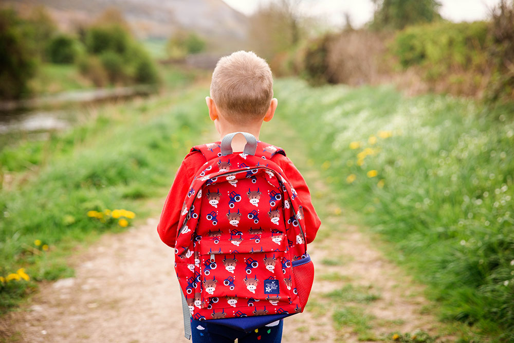 A boy in a red jumper wearing a red backpack with a cow and tractor pattern