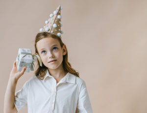 A young girl in a party hat shaking a gift by her ear