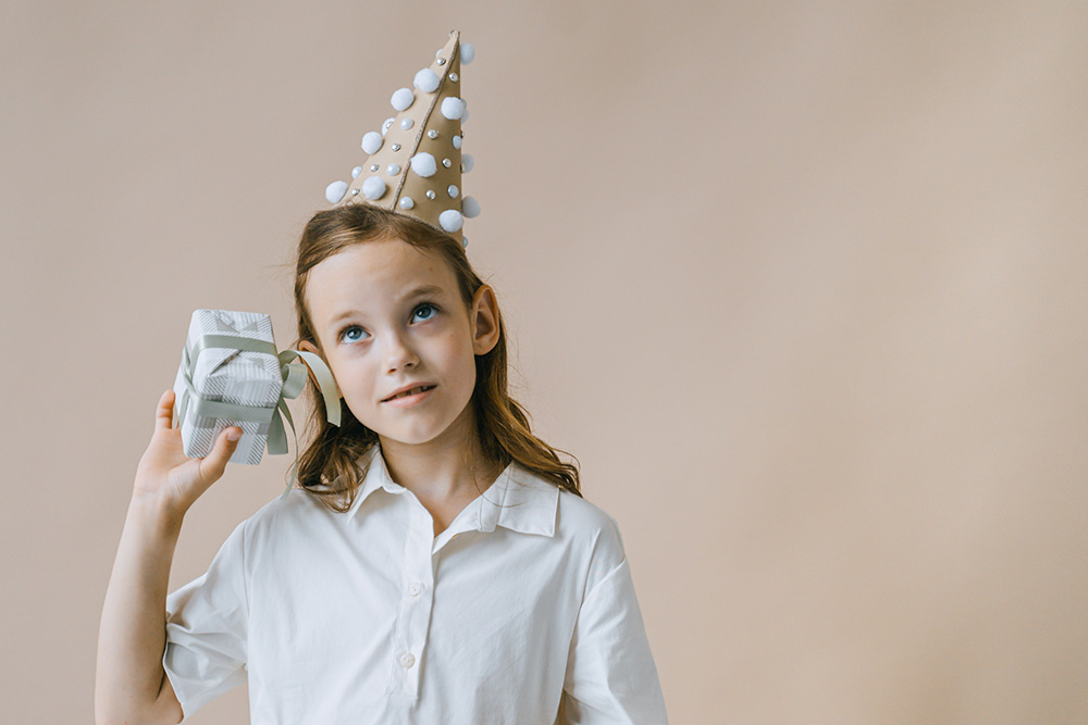 A young girl in a party hat shaking a gift by her ear