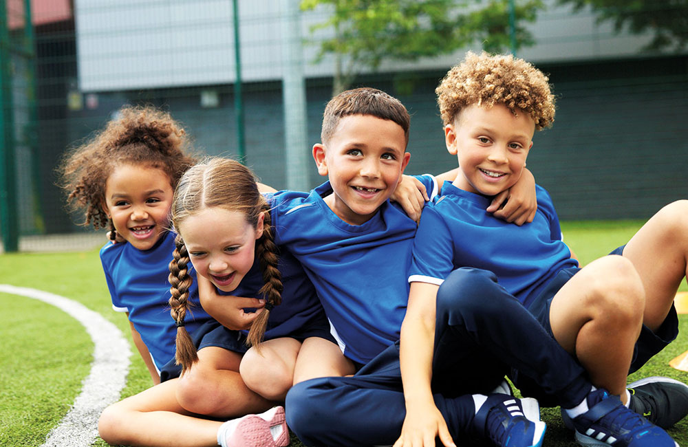 Four children sat on the floor outside wearing blue sports kits by Banner