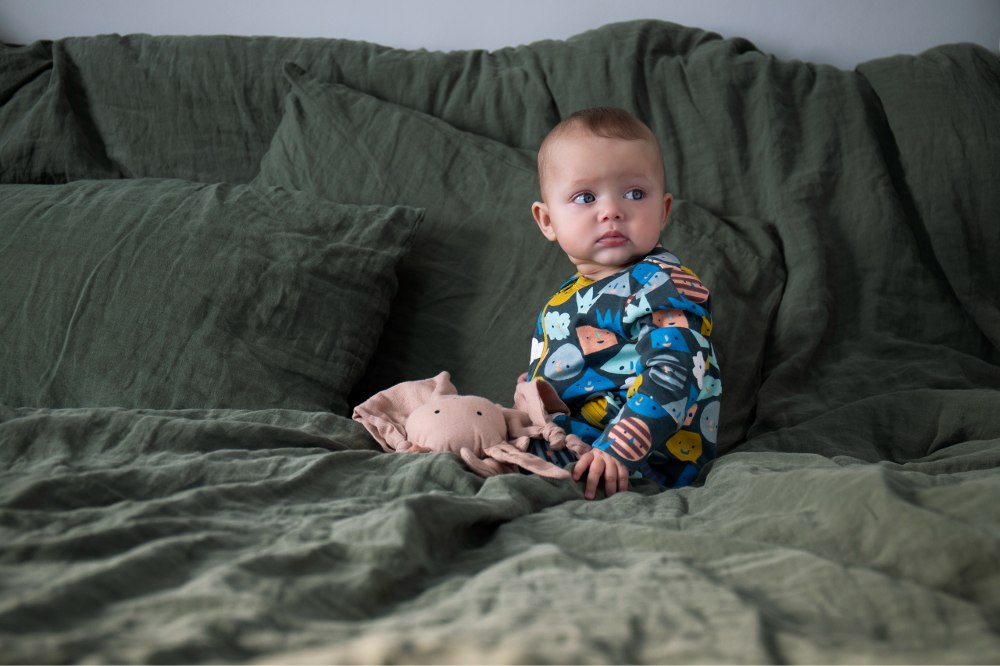 Baby wearing Kidly babygro sat on a bed with green bedding 