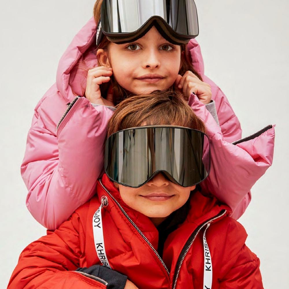 Girl stood in a pink jacket with ski goggles on her had behind a boy in a red jacket wearing ski goggles