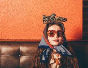 Girl sat on a leather set against an orange wall wearing sunglasses and a headscarf with a Zig + Star shoe on her head