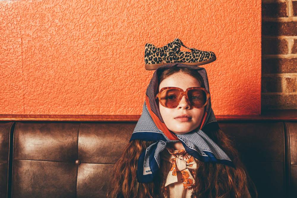 Girl sat on a leather set against an orange wall wearing sunglasses and a headscarf with a Zig + Star shoe on her head