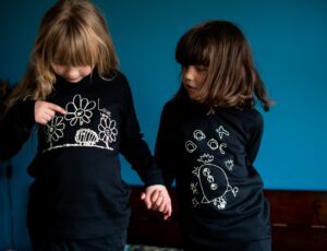 Two girls stood in a blue room wearing black pyjamas with prints of children's drawings on them