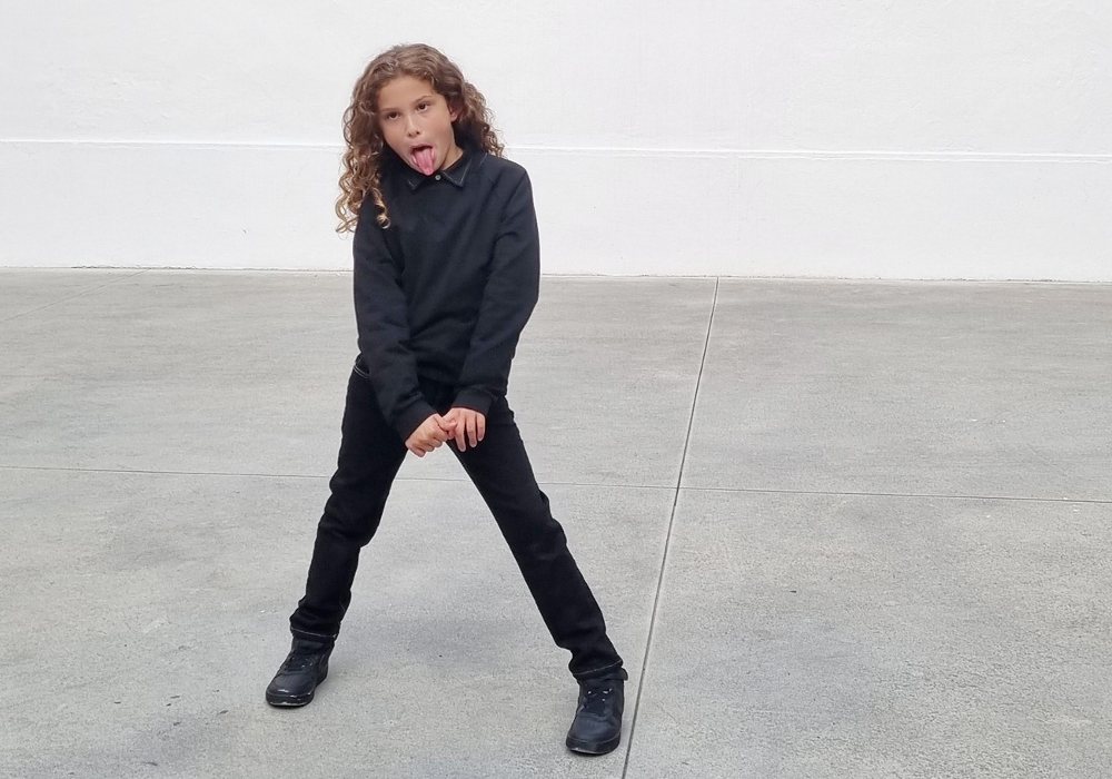 Girl stood on a grey floor against a white background wearing black trousers, a black top and black boots