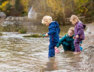 Three children stood in a river looking into the water wearing Kidunk outerwear