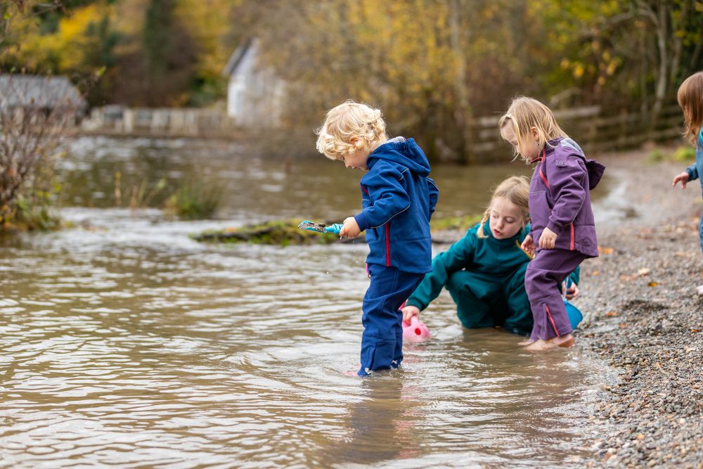 Three children stood in a river looking into the water wearing Kidunk outerwear