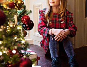 Girl sat on the floor looking at a Christmas tree wearing a red tartan Christmas dress by Lola Starr
