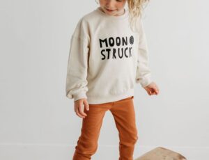 Girl stood balancing on a wooden bench wearing leggings and a white sweater with a MoonKids Collective logo