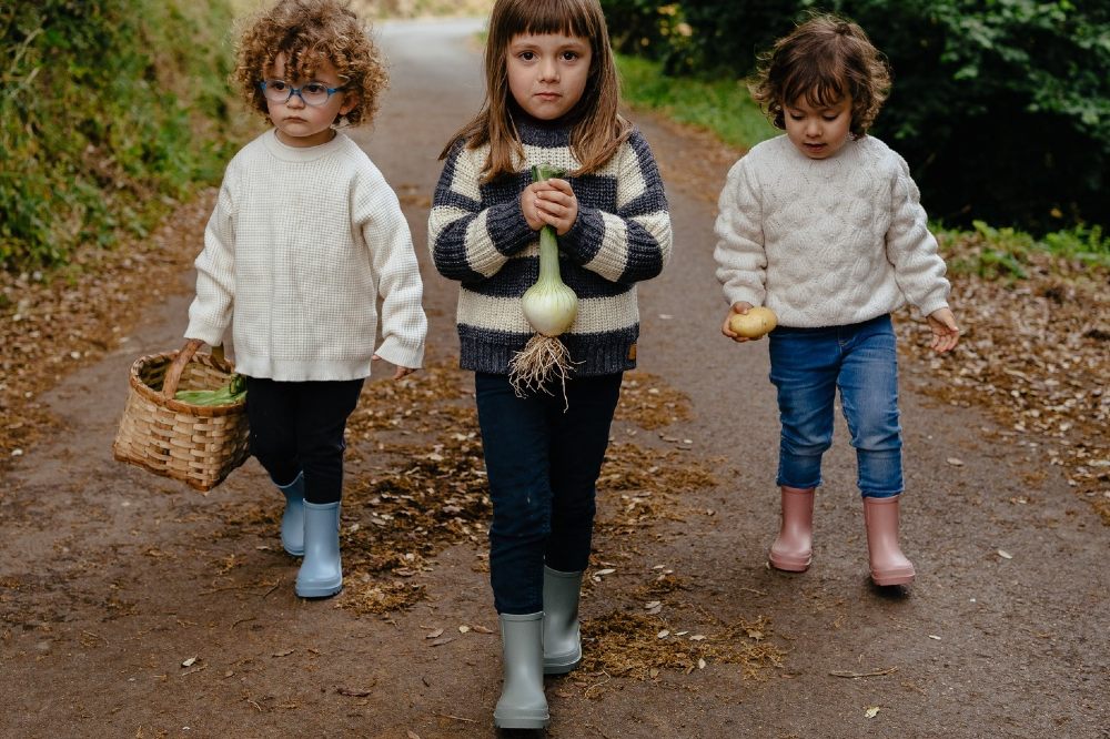Three children walking down a road wearing wellies and carrying a basket and vegetables