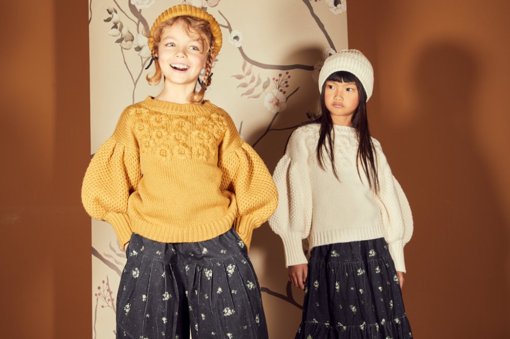 Two young girls stood against a patterned wall wearing wooly hats and jumpers
