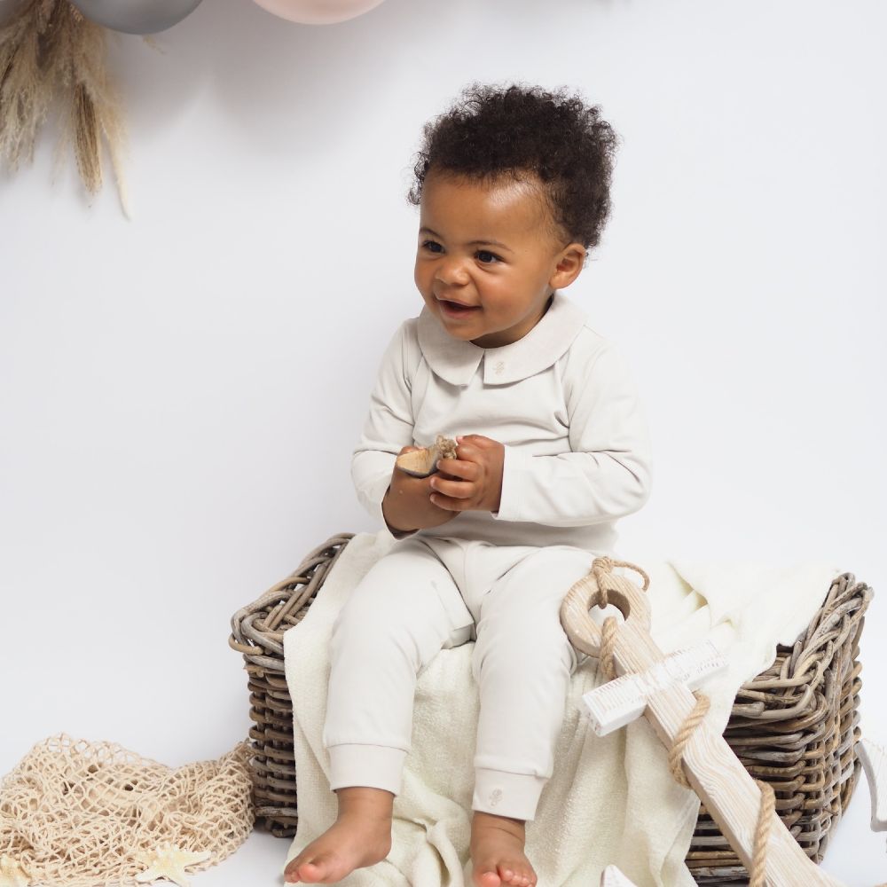 Toddler boy dressed in a white top and trousers sat on a wicker basket 