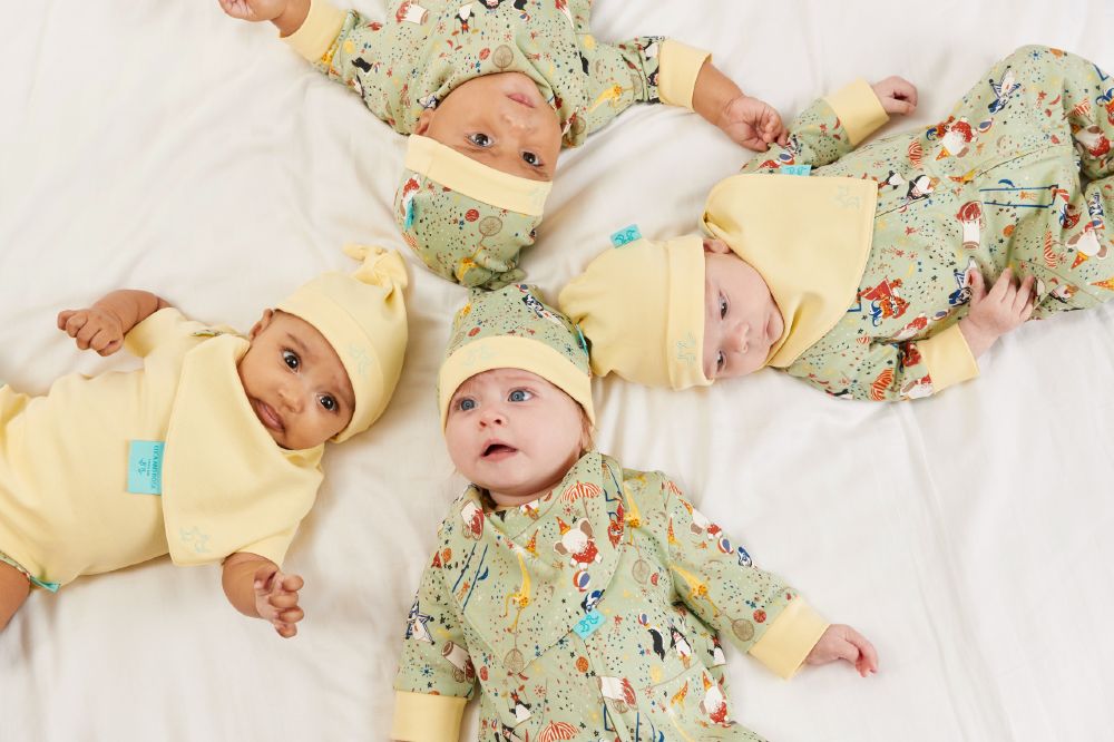Four babies lying on white bedding wearing hats and babygros by Luca and Rosa London