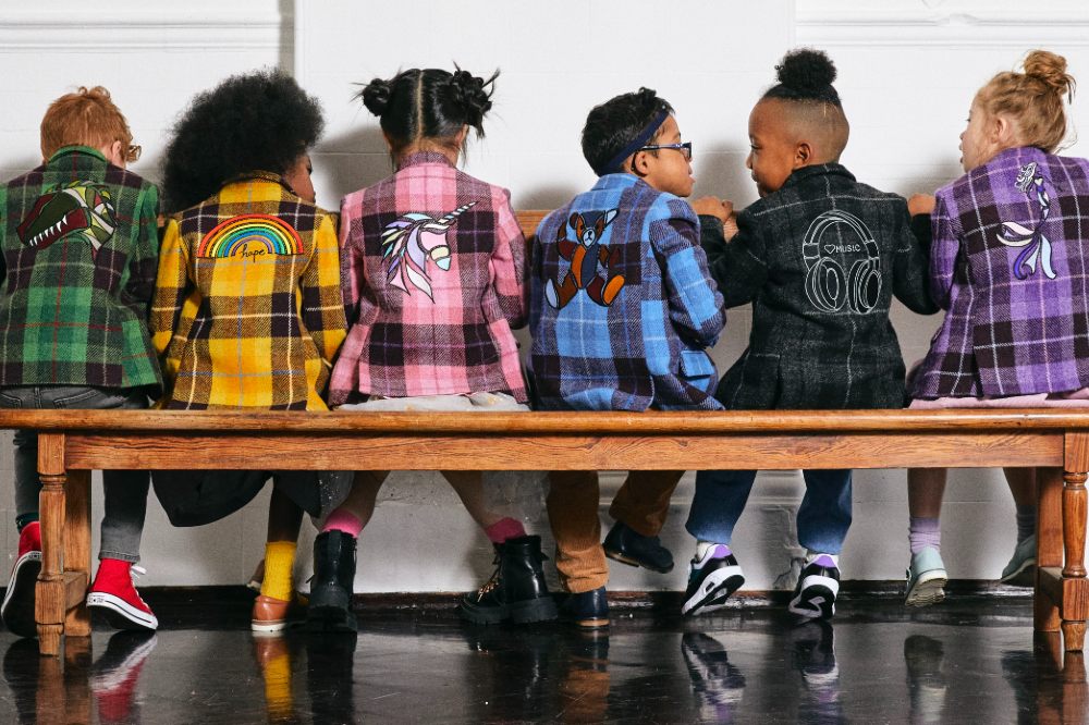 A row of children sat on a wooden bench with their backs to the camera wearing different coloured checked blazers 