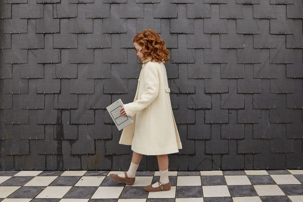 Girl with Red curly hair walked on a tiled floor wearing a cream coloured Twin & Chic coat