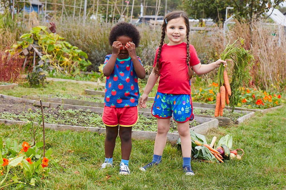 Two girls standing in an allotment holding tomatoes and a bunch of carrots