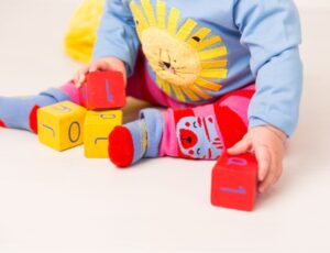 A baby sat on the floor playing with coloured wooden blocks wearing a top with a lion on and leggings by Blade & Rose