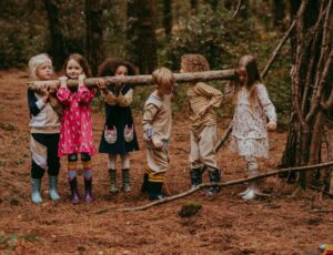 A group of children stood in a wood holding onto a tree branch wearing Lilly + Sid clothing
