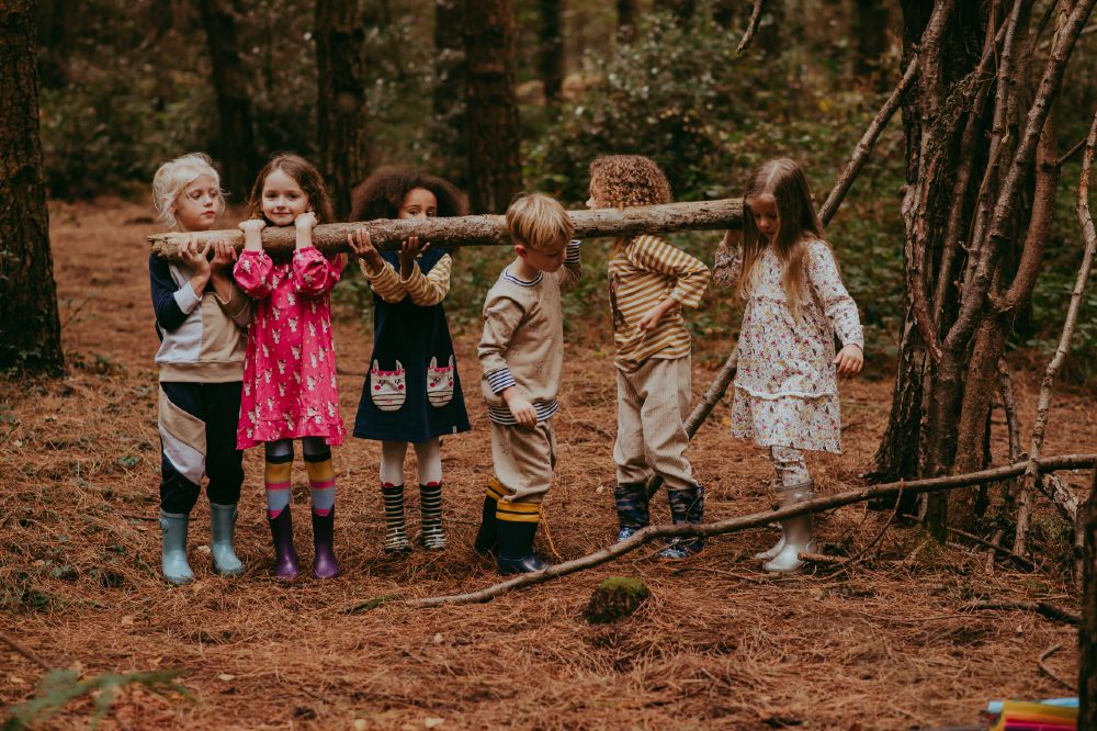 A group of children stood in a wood holding onto a tree branch wearing Lilly + Sid clothing