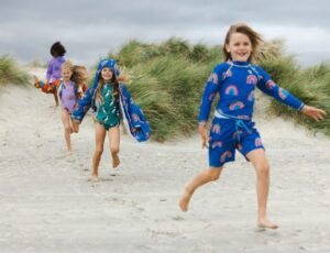 Children run down a beach wearing swimwear and outerwear from the Muddy Puddles Planet Happy Collection
