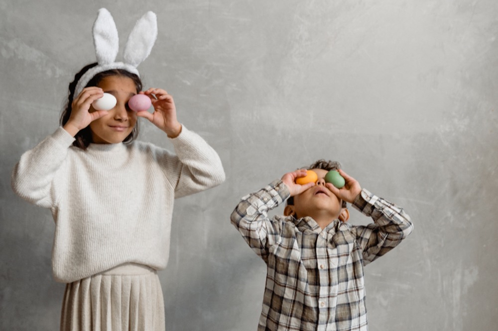 Two children holding easter eggs up to their eyes with one of the children wearing bunny ears